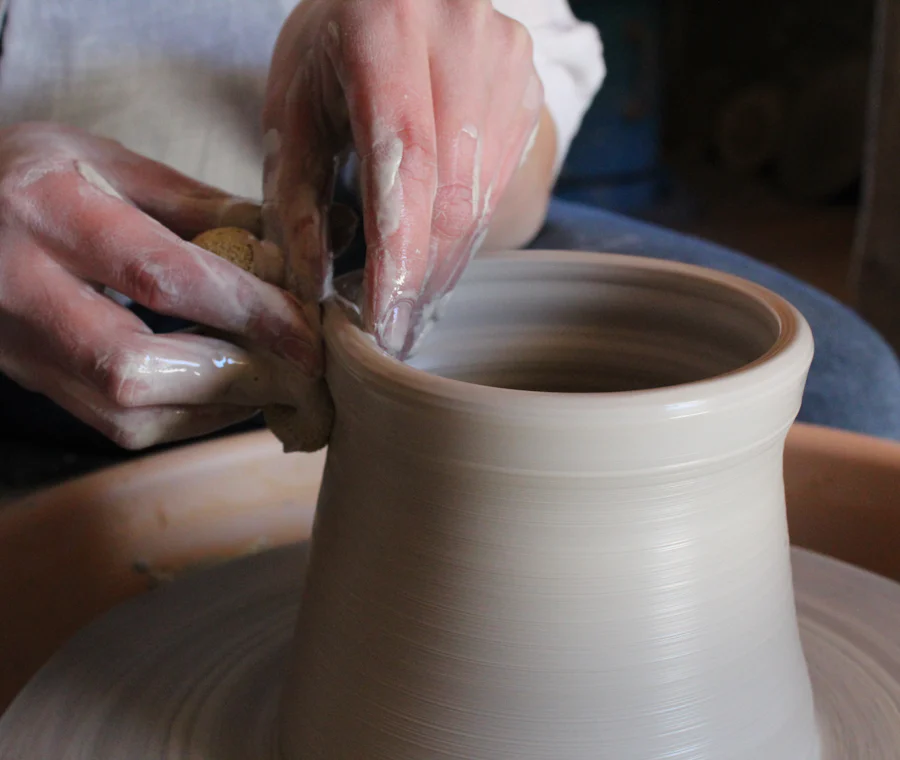 A potter's clay-covered hands shaping the rim of a vessel on a turning wheel.